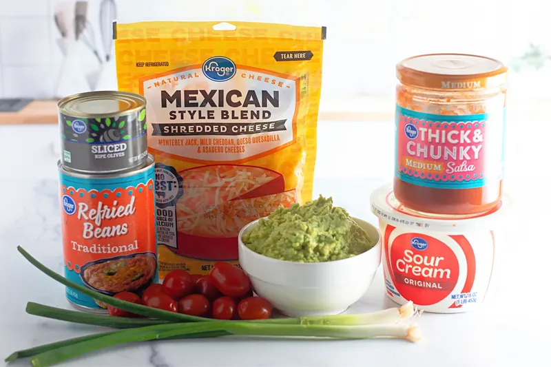 Ingredients for 7 layer dip cups including refried beans, guacamole, cheese, salsa, sour cream and toppings