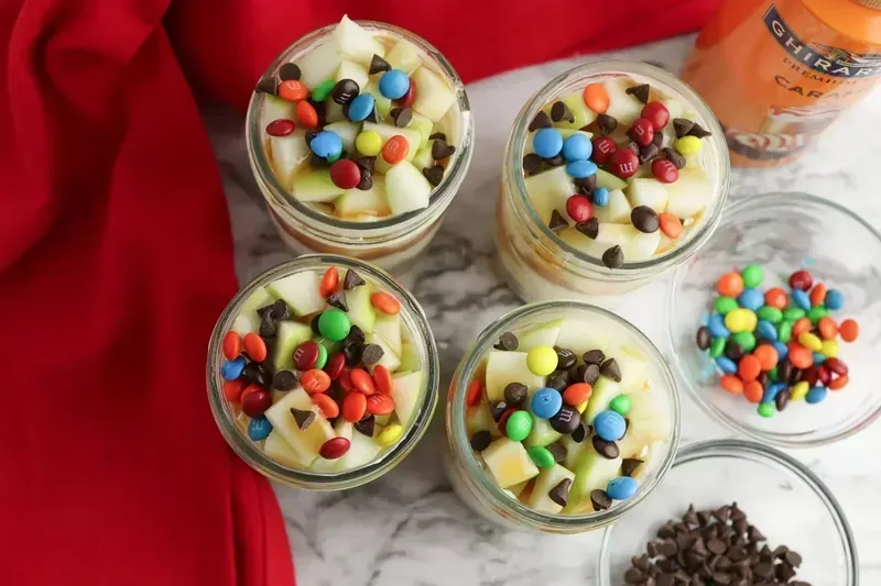 Cheesecake jars topped with colorful mini M&Ms and chocolate chips over caramel-drizzled apples