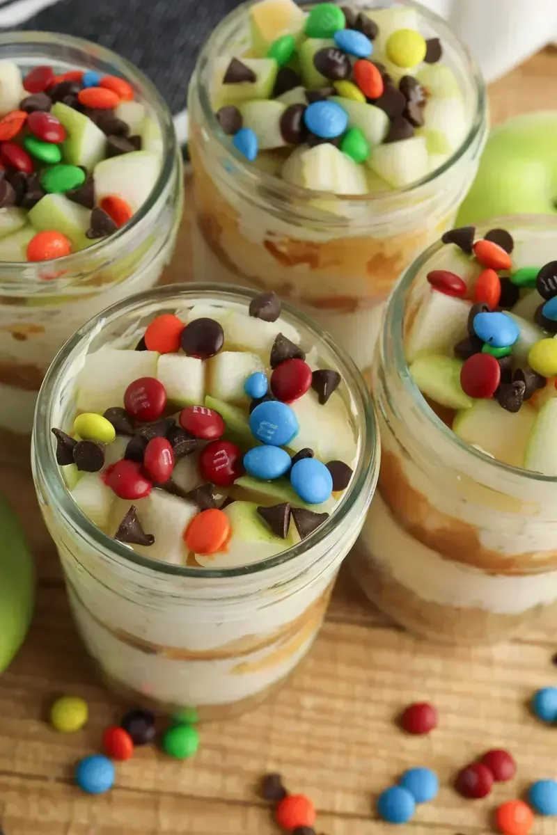 Four candy apple cheesecake jars topped with diced green apples, mini M&Ms, and chocolate chips