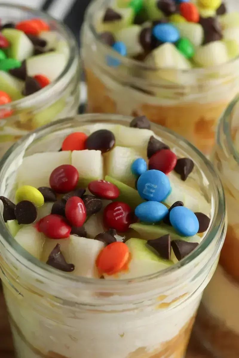 Candy apple cheesecake jars with creamy layers, caramel sauce, diced apples, and colorful M&Ms and chocolate chips