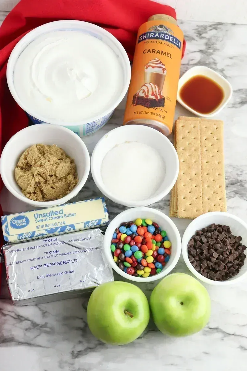 Ingredients for candy apple cheesecake jars including cream cheese, caramel sauce, graham crackers, apples, M&Ms, and chocolate chips