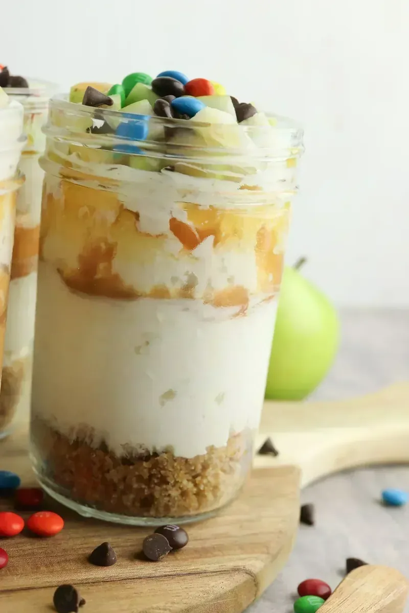 Completed candy apple cheesecake jars displayed on wooden surface with scattered candies and fresh apple