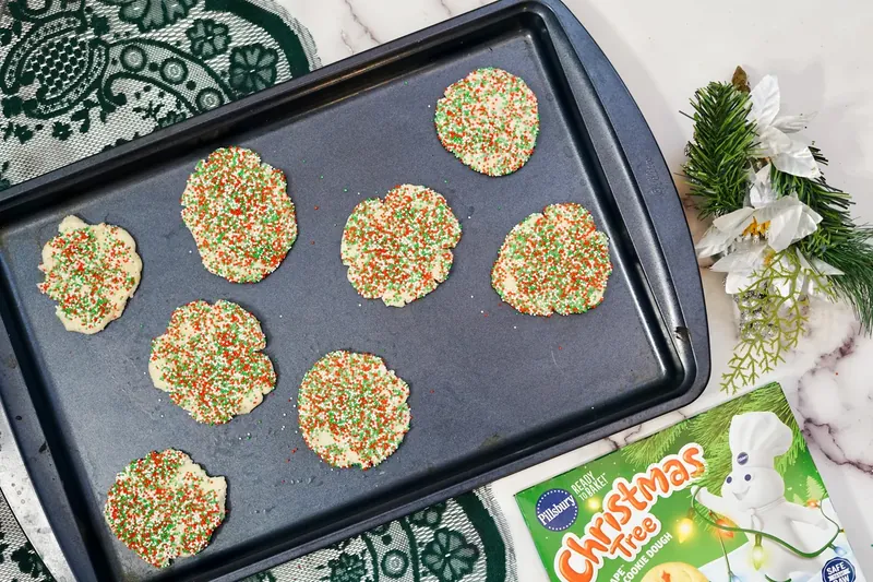 Flattened sugar cookies covered in Christmas sprinkles arranged on dark baking sheet