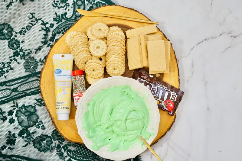 Ingredients for Christmas tree frosting board including green frosting, cookies, graham crackers, M&M candies, and yellow icing