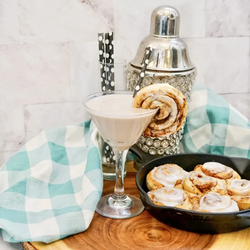 Elegant cinnamon roll martini with cinnamon roll garnish and fresh cinnamon rolls in background
