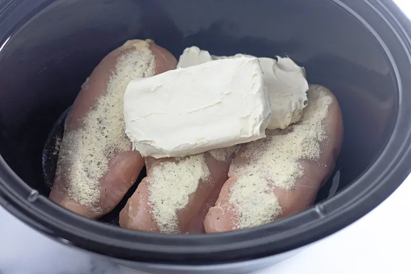 Raw chicken breasts with ranch seasoning and cream cheese blocks in slow cooker