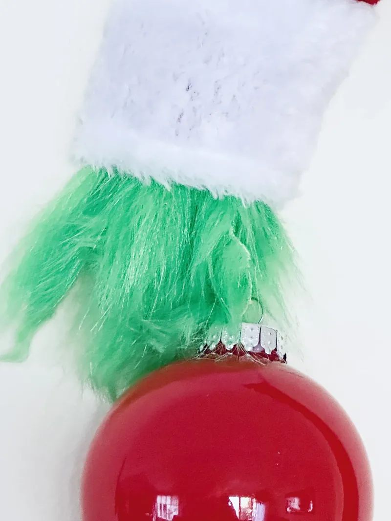 Close up of completed Grinch hand with green fur fingers holding red Christmas ornament