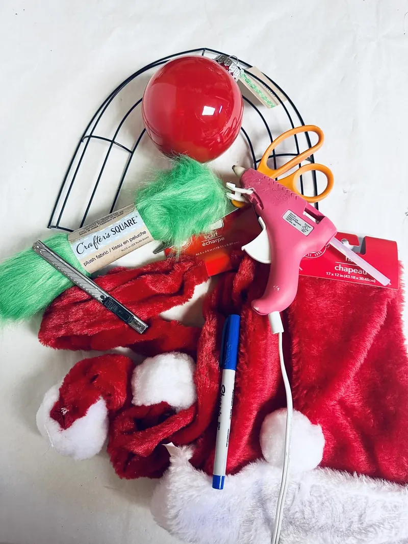 Craft supplies for Grinch wreath including candy cane wreath form, red scarf, green faux fur, red ornament, hot glue gun and scissors