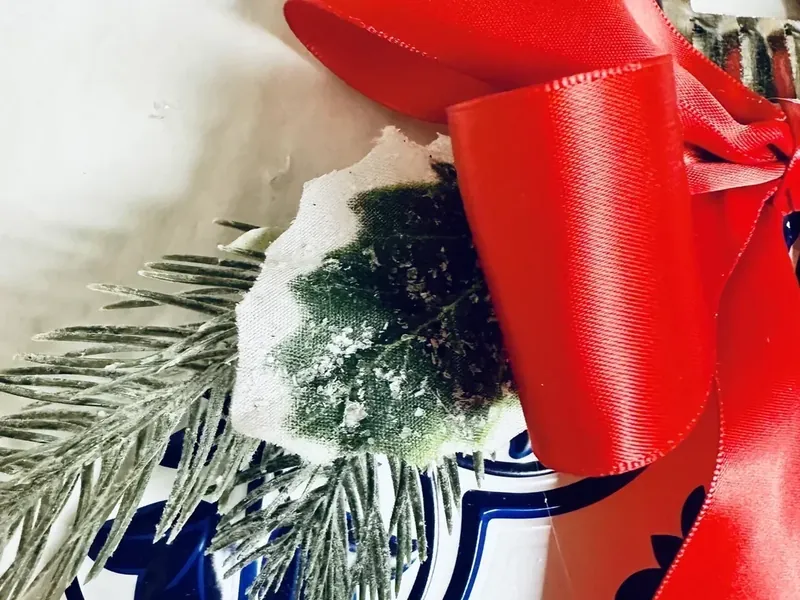 Artificial pine branches and holly being glued to ornament wreath with red ribbon bow