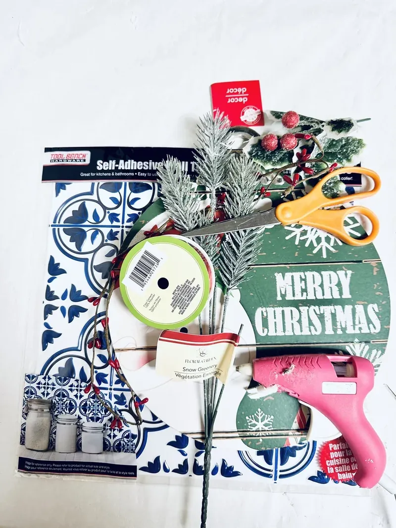 Dollar Tree craft supplies including blue and white tile, ornament sign, pine picks, ribbon, and hot glue gun