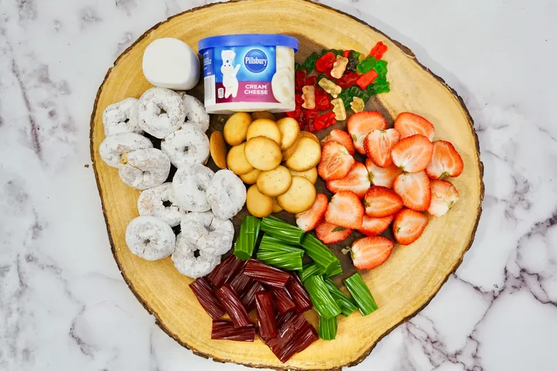 Raw ingredients for gnome frosting board including vanilla frosting, sliced strawberries, marshmallow, vanilla wafers, and assorted treats