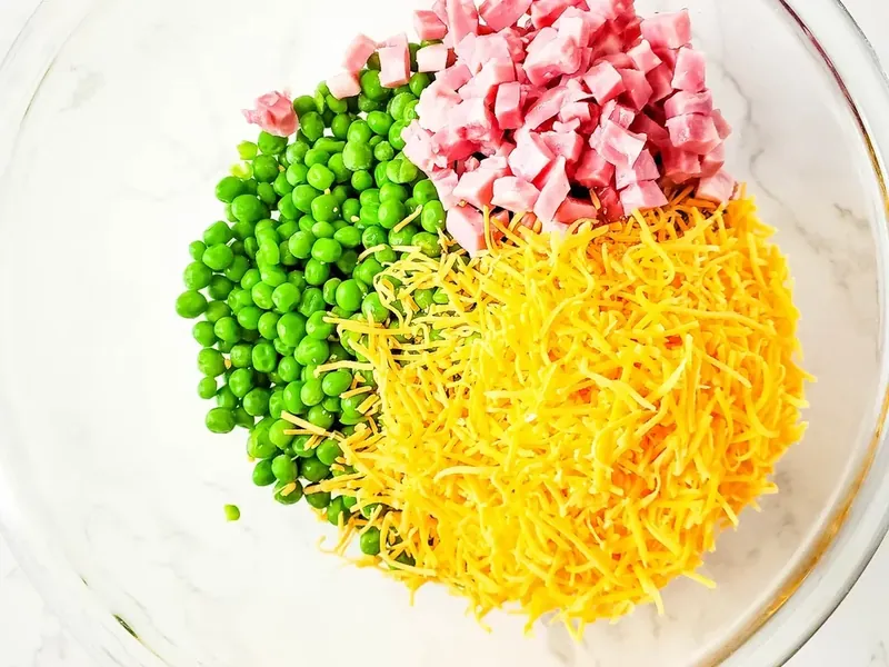 Shredded cheddar cheese being added to peas and ham in bowl