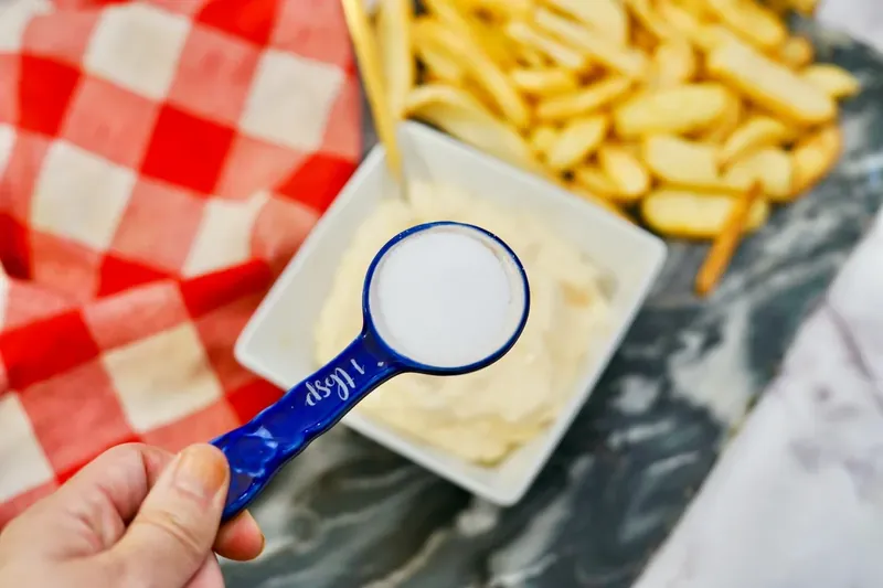 Hand holding blue measuring spoon with sugar over ingredients for In-N-Out sauce