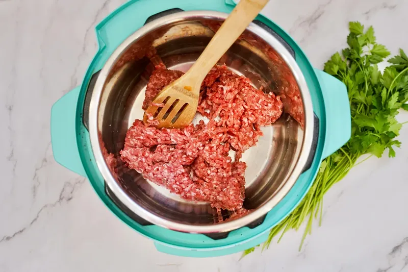 Raw ground Italian sausage being browned in Instant Pot with wooden spoon