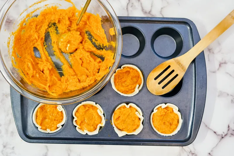 Pumpkin filling being spooned into pie crusts in muffin tin