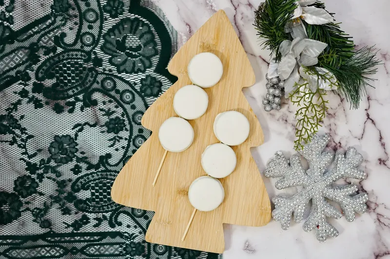 Three white chocolate covered Oreos threaded on wooden skewers on Christmas tree cutting board