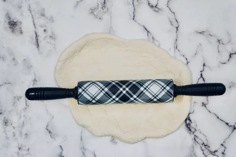 Pizza dough rolled flat with decorative plaid rolling pin on marble countertop for pepperoni rose balls recipe