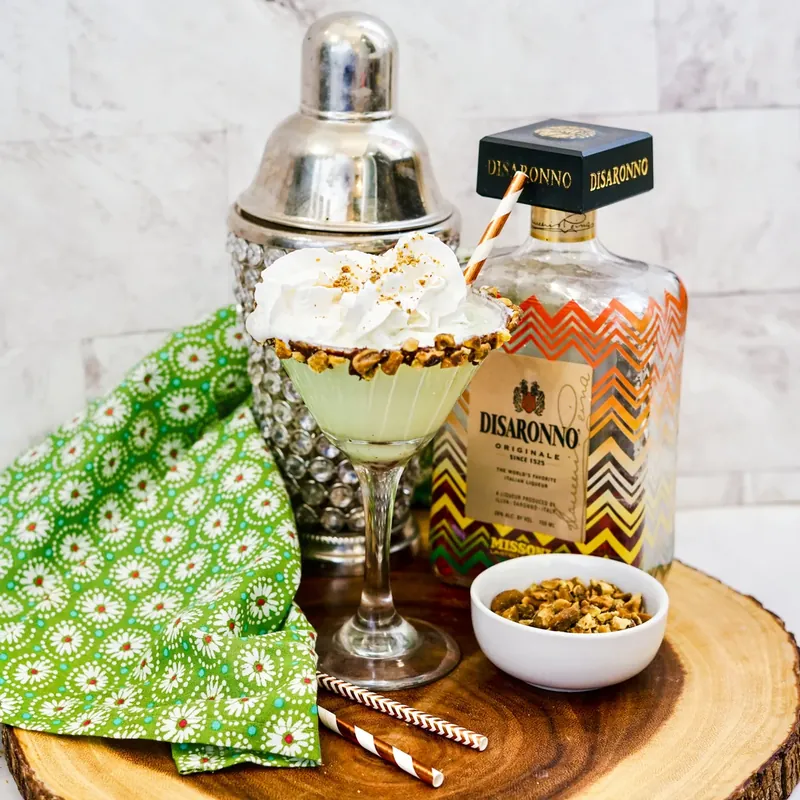 Pistachio martini styled with cocktail shaker and Disaronno bottle on wooden board
