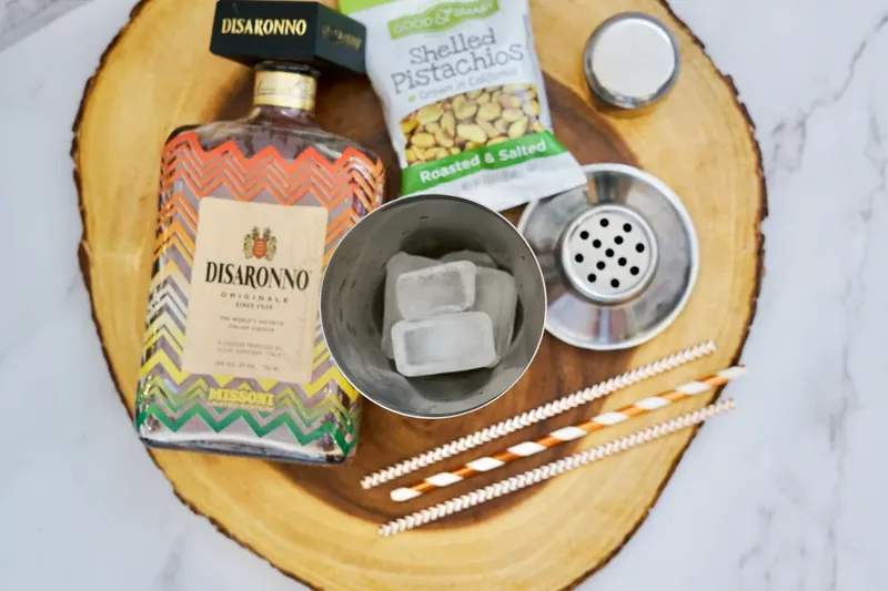 Ice cubes in cocktail shaker with ingredients and pistachios on wooden board