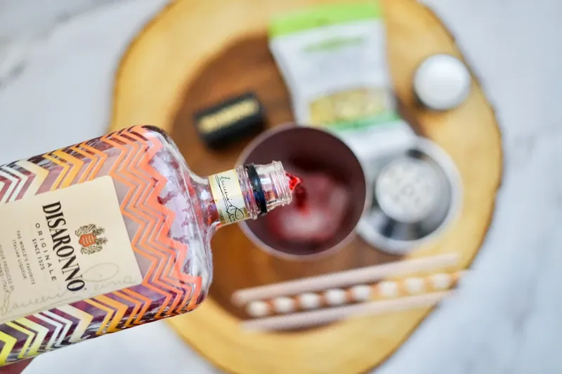 Pouring Disaronno liqueur into cocktail shaker with other ingredients on wooden board
