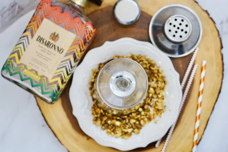 Martini glass being rolled in crushed pistachios on plate with Disaronno bottle