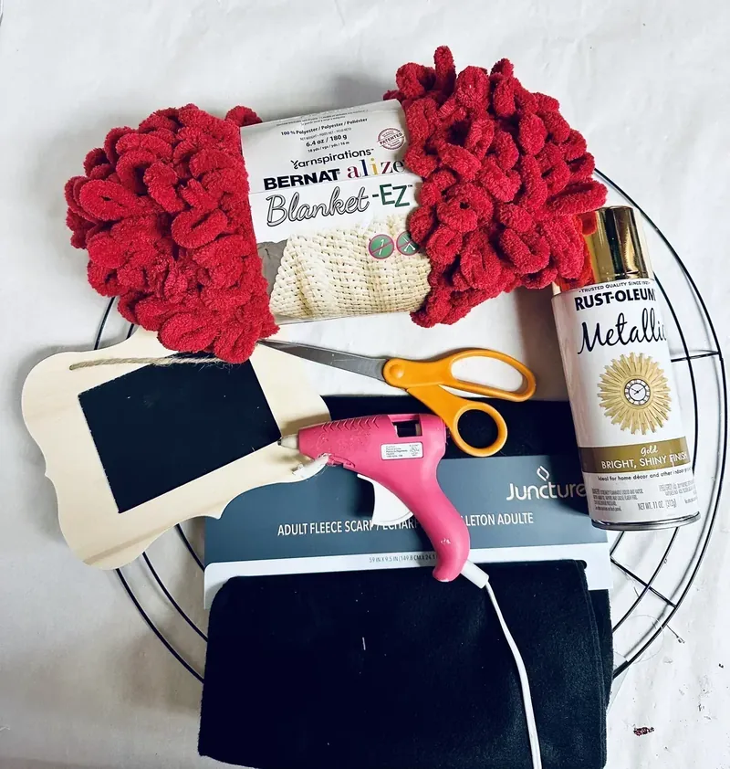 Craft supplies for Santa belt wreath including red Bernat Alize yarn, wire wreath form, black fleece scarf, wooden chalkboard frame, gold spray paint, hot glue gun and scissors