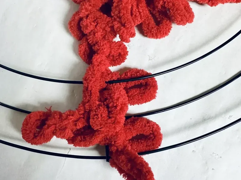 Red textured yarn being tied to black wire wreath form to begin wrapping process