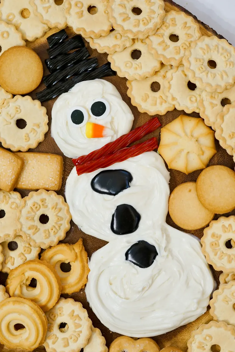 Snowman frosting board with white frosting snowman, candy eyes, candy corn nose, and assorted cookies on wooden tray