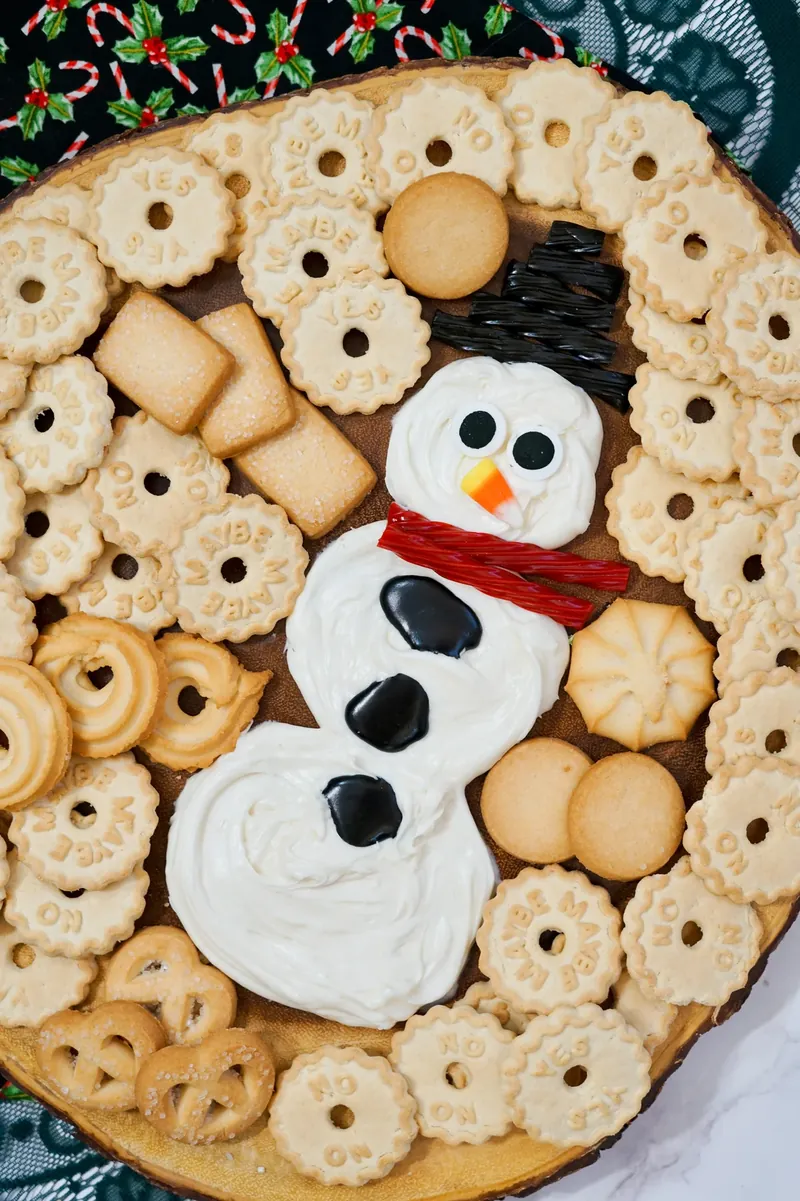 Complete snowman frosting board with white frosting snowman, black hat, red scarf, and variety of cookies arranged on round wooden board