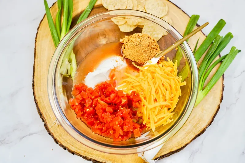 Glass bowl with sour cream, shredded cheddar cheese, RoTel, and taco seasoning being mixed together
