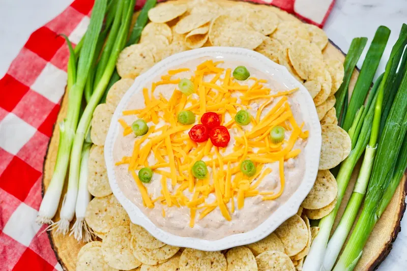 Finished TikTok boat dip in white serving bowl garnished with cheddar cheese and green onions, surrounded by tortilla chips