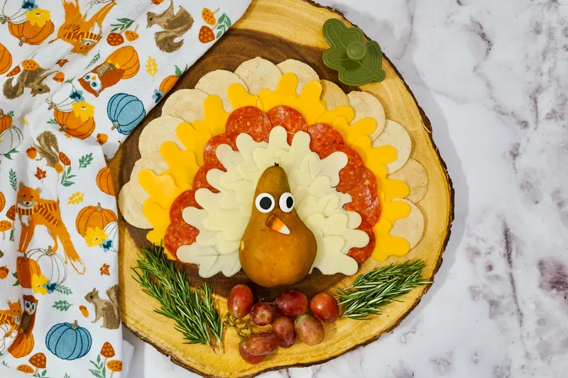 Creative Thanksgiving turkey cheese board with arranged cheese and pepperoni feathers, pear turkey body, rosemary garnish and grapes