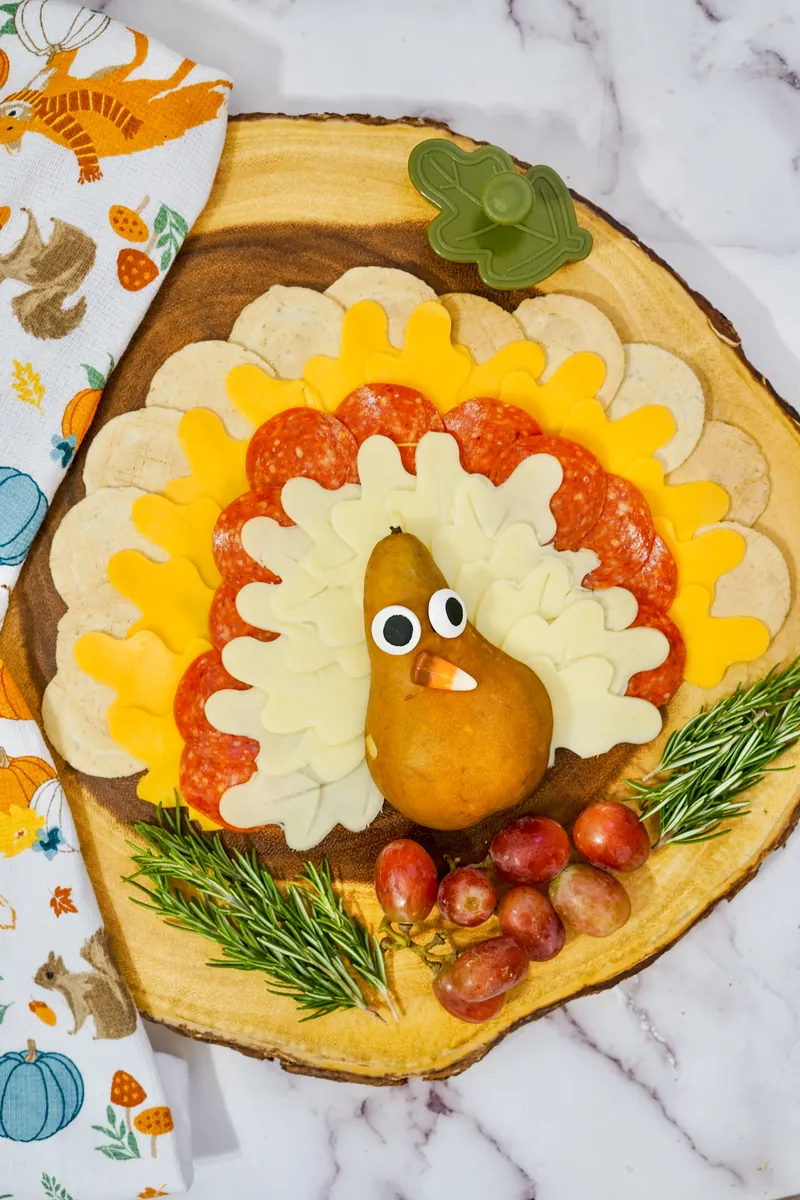 Turkey-shaped cheese platter with layered white and yellow cheese feathers, pepperoni, pear turkey body with googly eyes on rustic wood