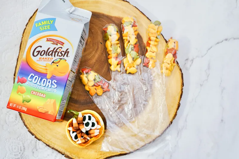 Clear plastic glove filled with colorful Goldfish crackers in fingers on wooden cutting board next to Goldfish box