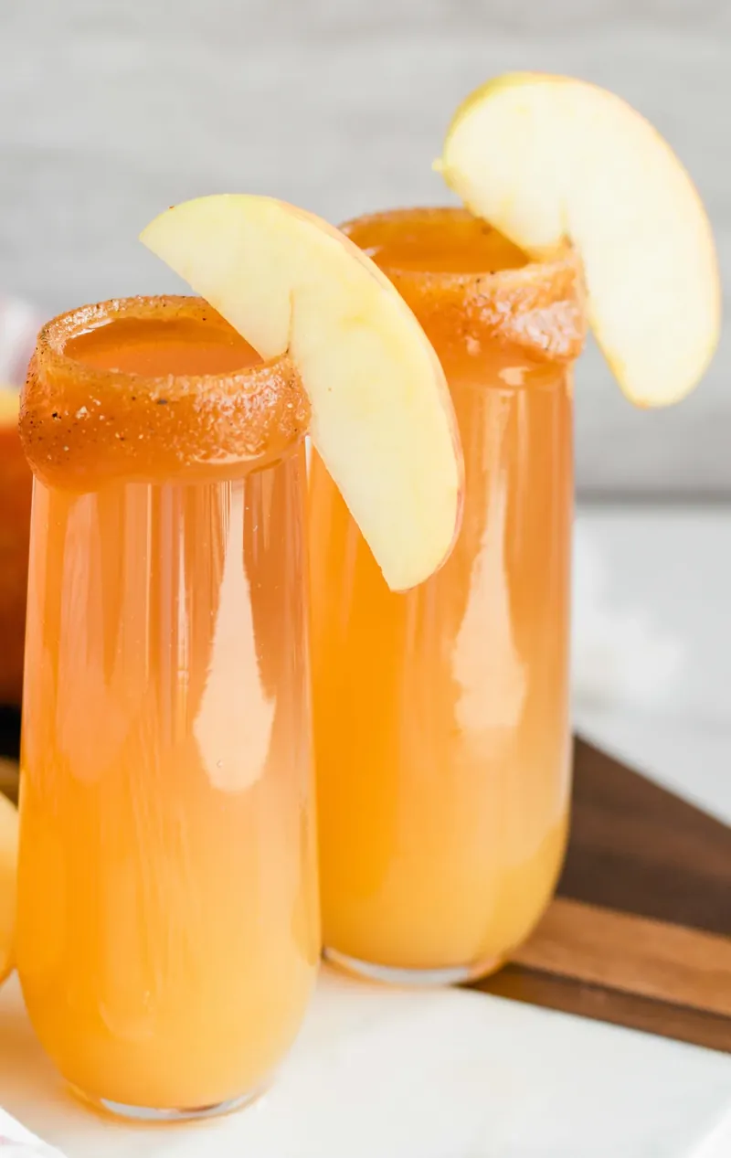 Two apple cider mimosas with caramel rim and apple slice garnish on wooden serving tray