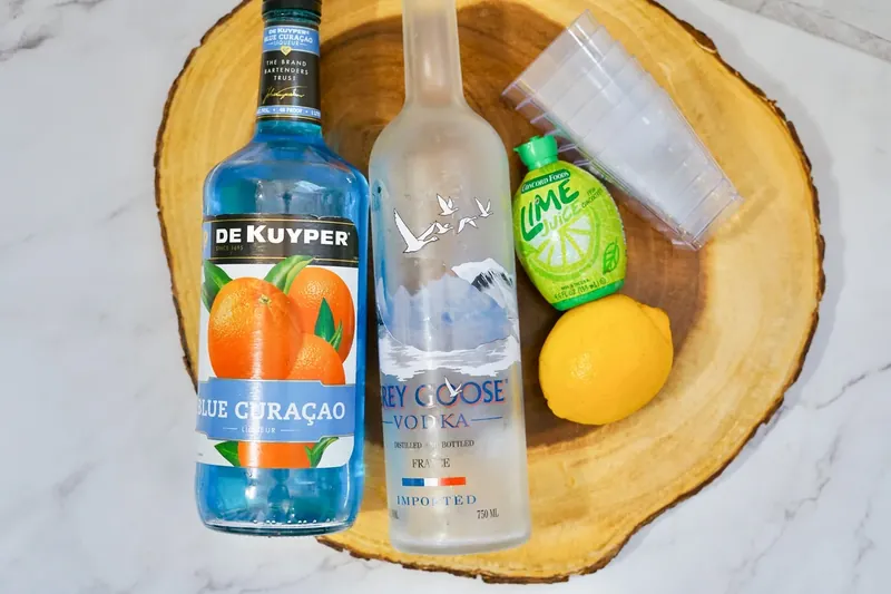 Blue kamikaze shooter ingredients including blue curacao, vodka, lime juice and lemon on wooden board