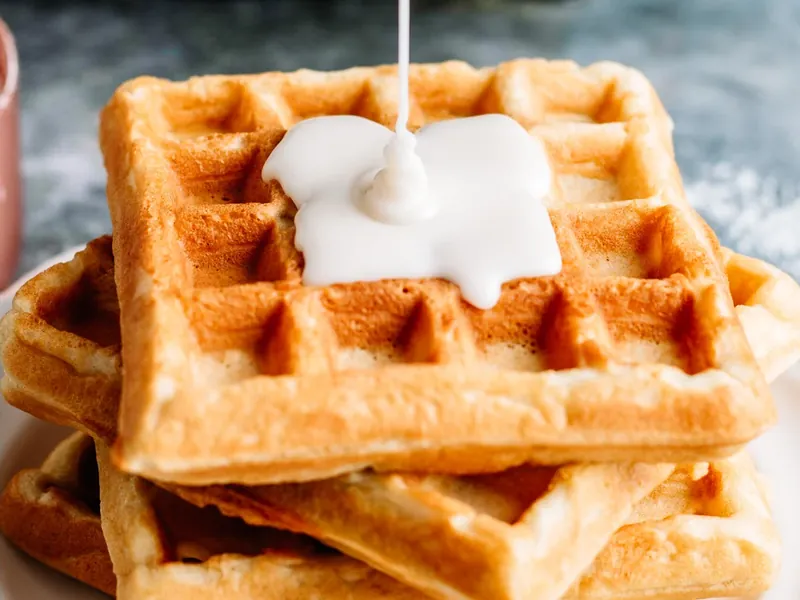 White glaze being drizzled over golden brown waffle from small bowl