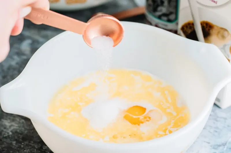 Hand adding salt to white bowl with milk egg butter and vanilla being mixed