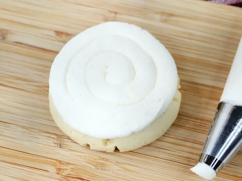 Piping bag creating spiral of white peppermint frosting on top of baked cookie on cutting board