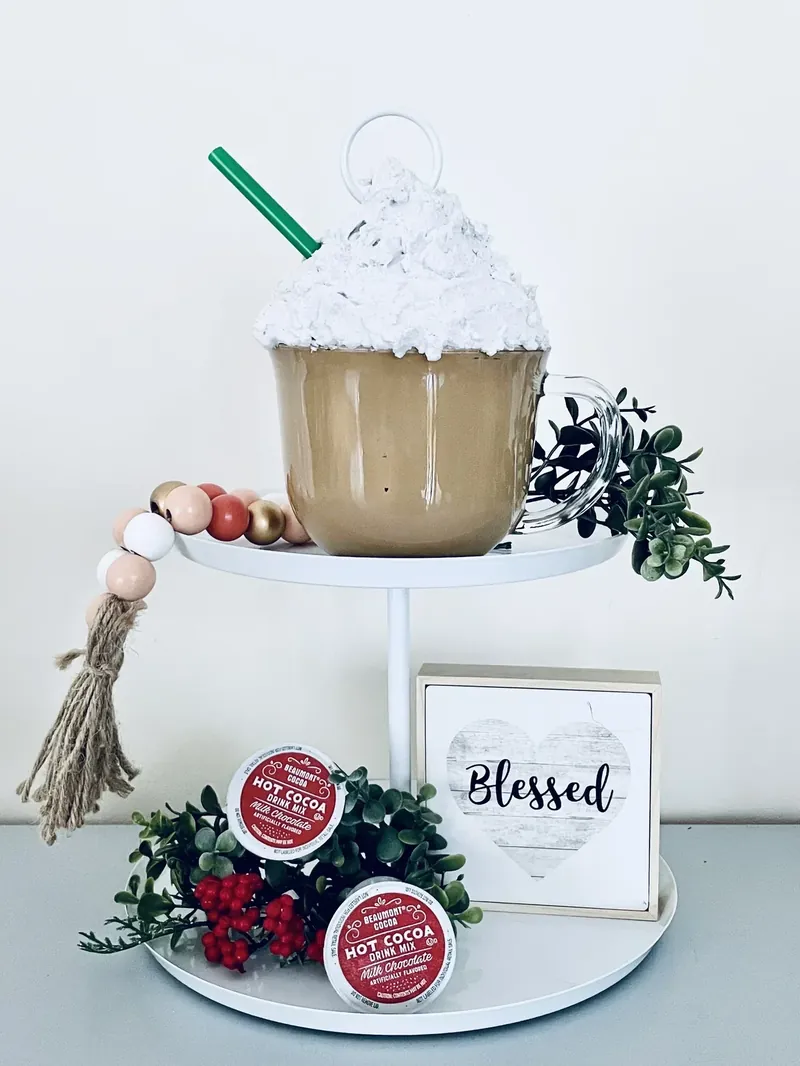 Completed Dollar Tree faux hot cocoa mug displayed on white tiered stand with holiday decorations and blessed sign