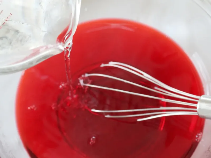 Pouring liquid into red jello mixture while whisking in glass bowl