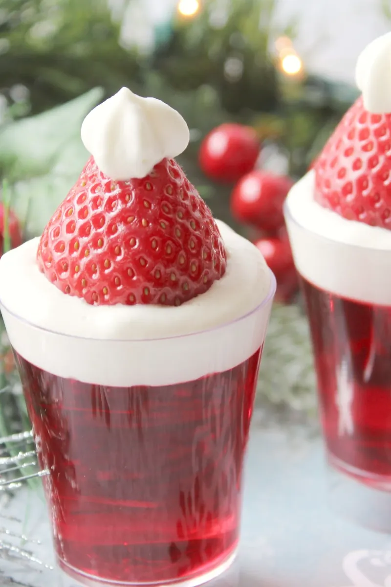 Santa hat jello shots with red jello, whipped cream and strawberry garnish on Christmas background
