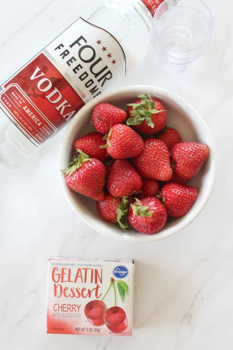 Ingredients for santa hat jello shots including vodka, fresh strawberries and cherry gelatin