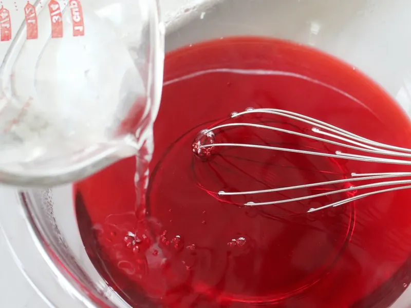 Whisking red jello mixture with vodka and water in glass measuring cup