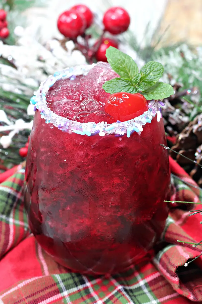Sugar plum cocktail with sparkling sugar rim and mint garnish surrounded by Christmas decorations
