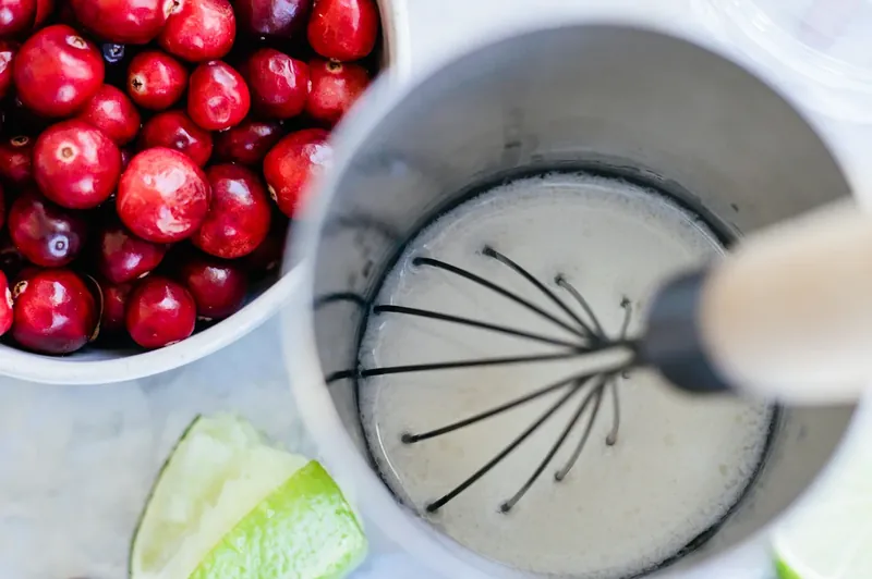 Whisking creamy white margarita mixture in bowl with cranberries and lime nearby