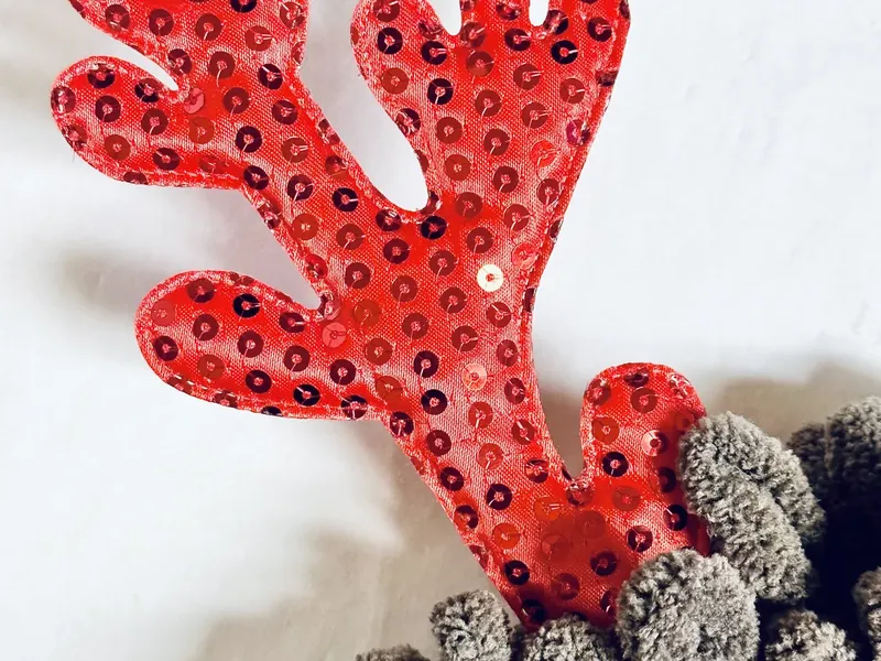 Red Christmas ornament being attached as nose to yarn-wrapped wreath with hot glue